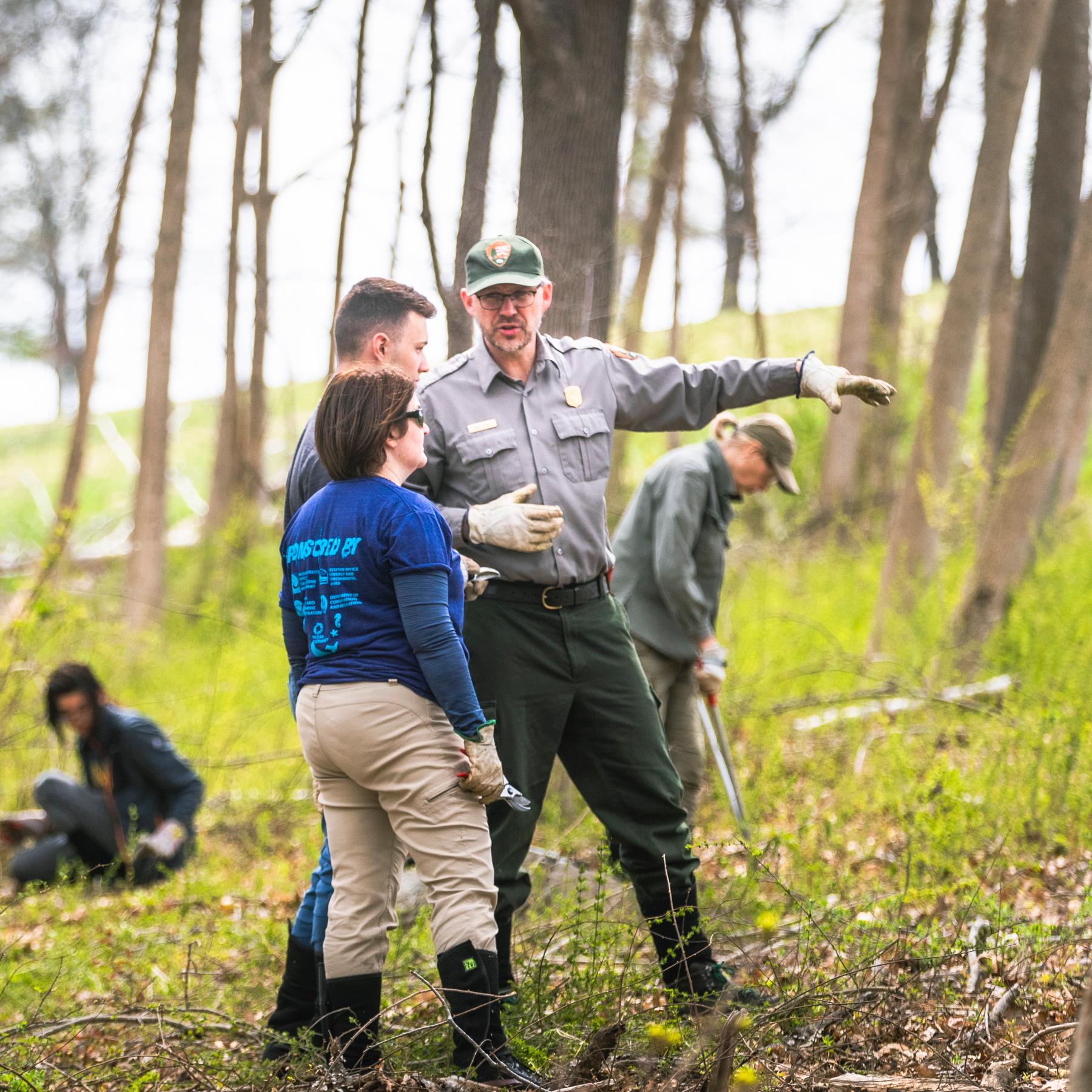 A uniformed ranger speaks to a group of volunteers standing in a grassy wooded area. 