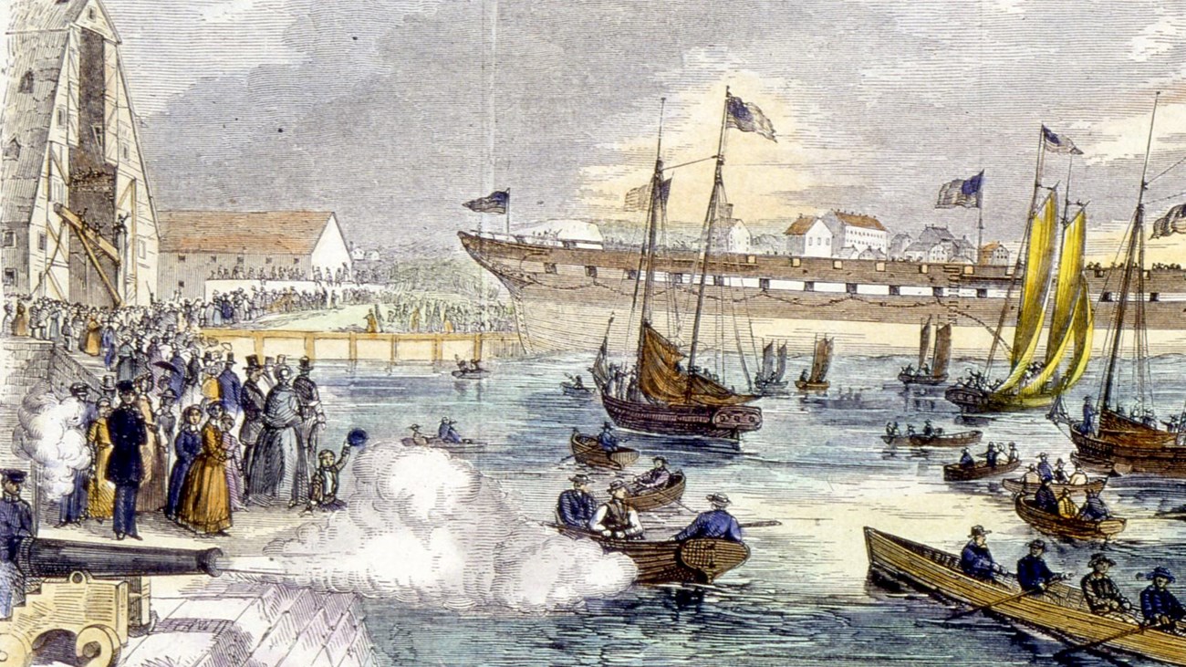 Hull of a ship launching into a harbor as crowds watch on piers and a swarm of boats. A cannon fires