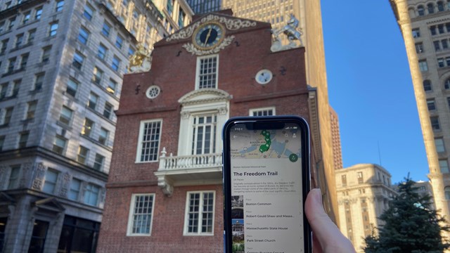 A hand holding up a phone with the NPS App open and in the background the Old State House.