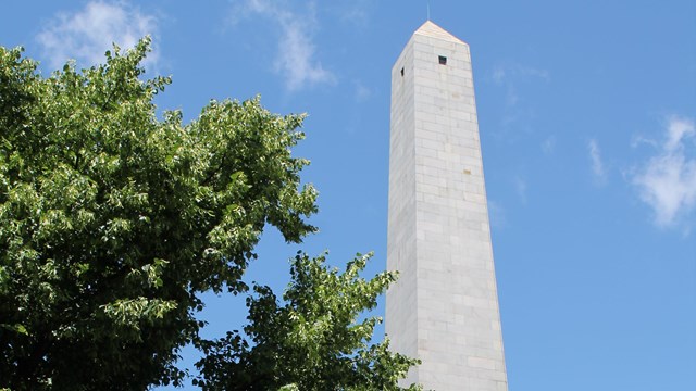 A white obelisk, the Bunker Hill Monument, against a blue sky with a green tree in the foreground.