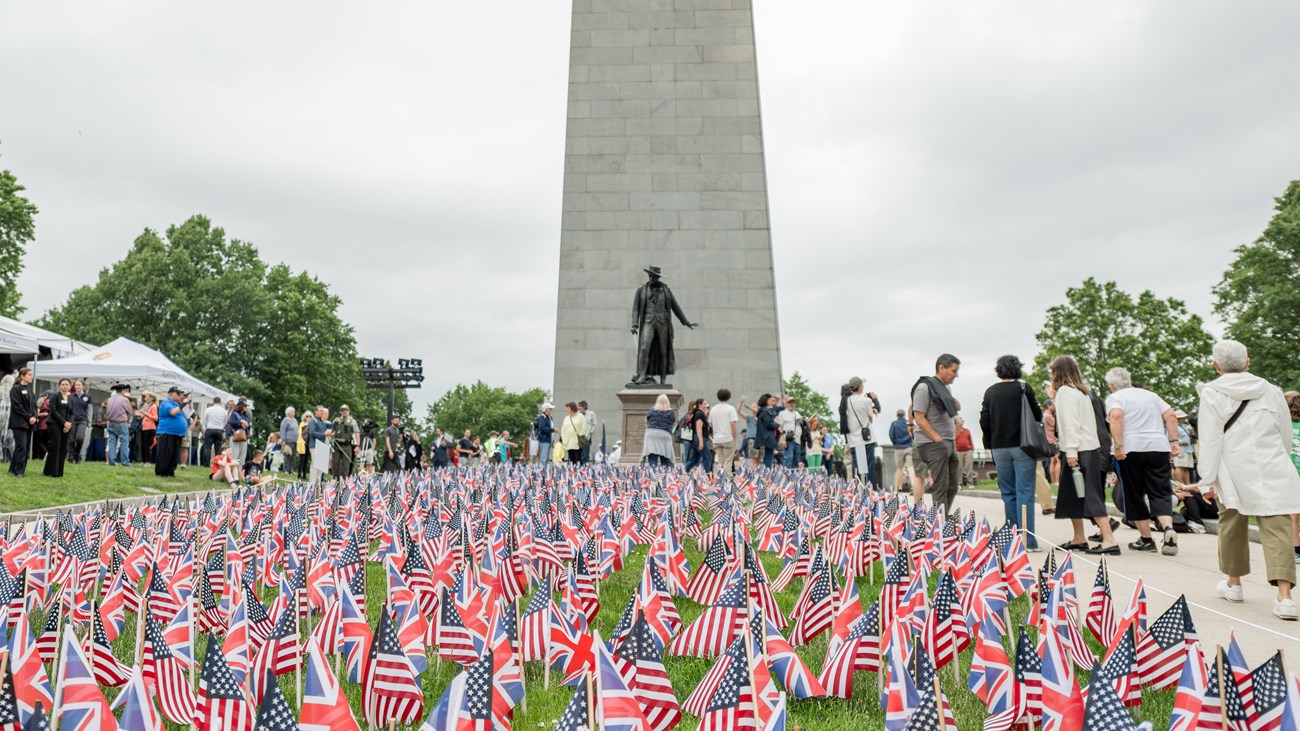 People surround a flag garden of American and British flags in front of the Bunker Hill Monument