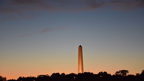 Photograph of a twilight sky with an illuminated obelisk in the center.