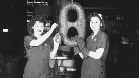 Photograph of two female workers displaying a small piece and a very large piece of anchor chain