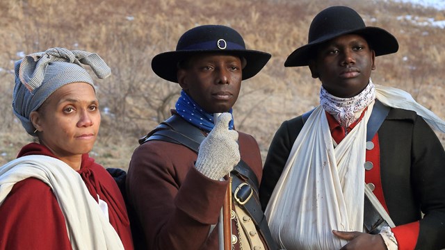 Three African American Revolutionary War reenactors, two men stand with a woman to the left. 