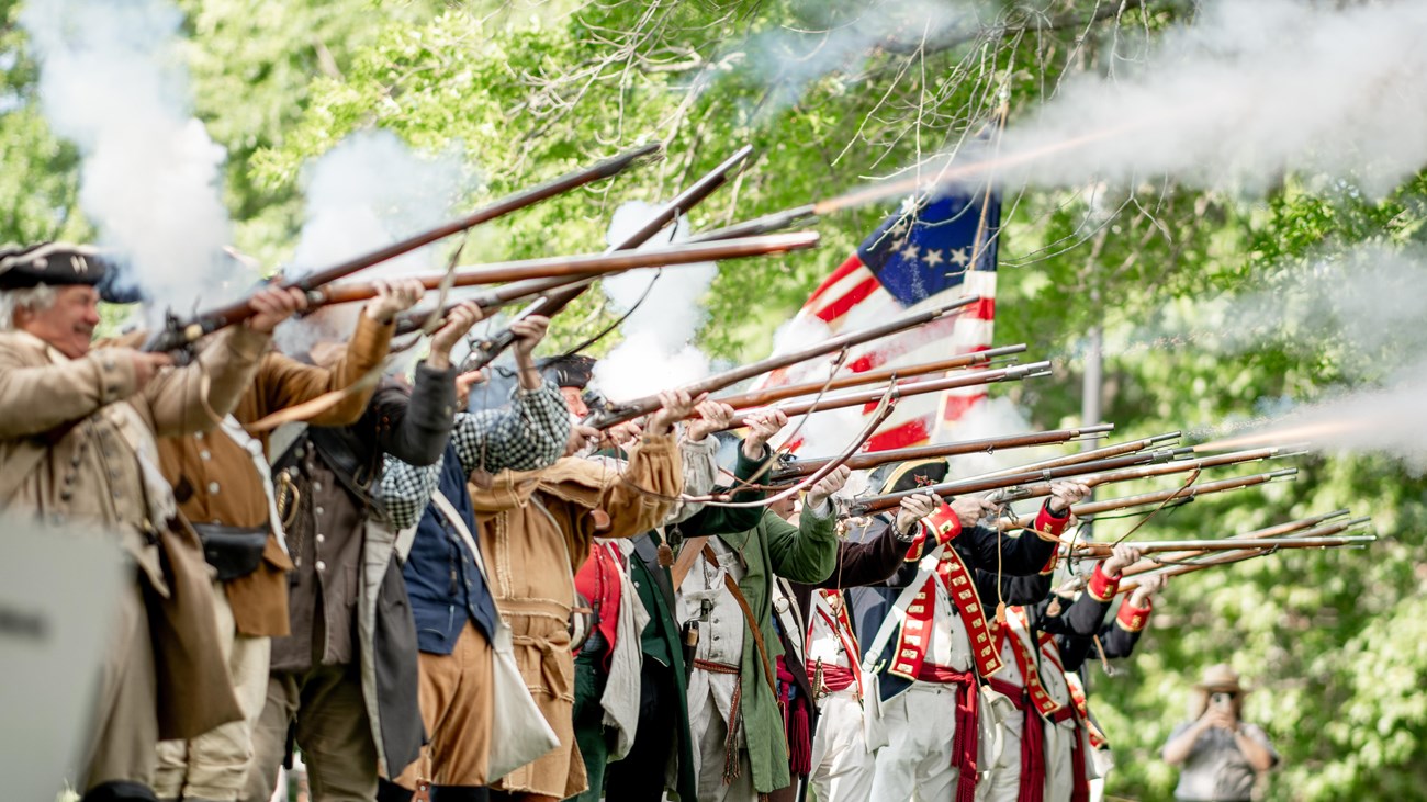 Men in colonial cloths and solider uniforms firing muskets.