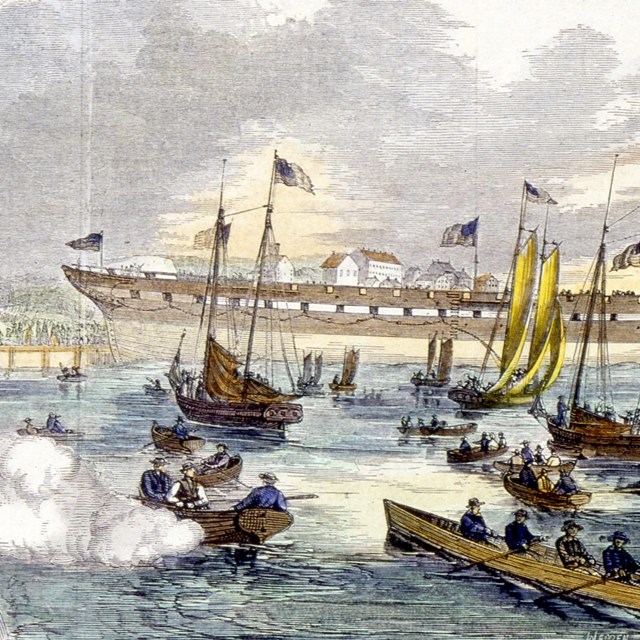 Hull of a ship launching into a harbor as crowds watch on piers and a swarm of boats. A cannon fires