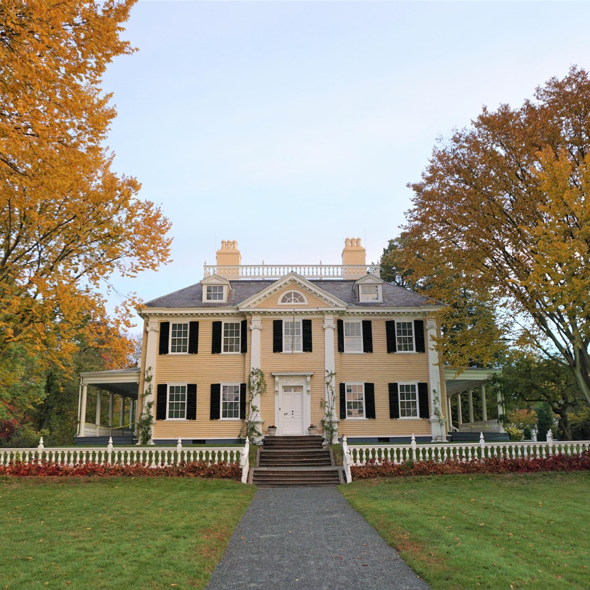 Facade of 3-story symmetrical yellow Georgian house with large green front lawn, flanked by trees.