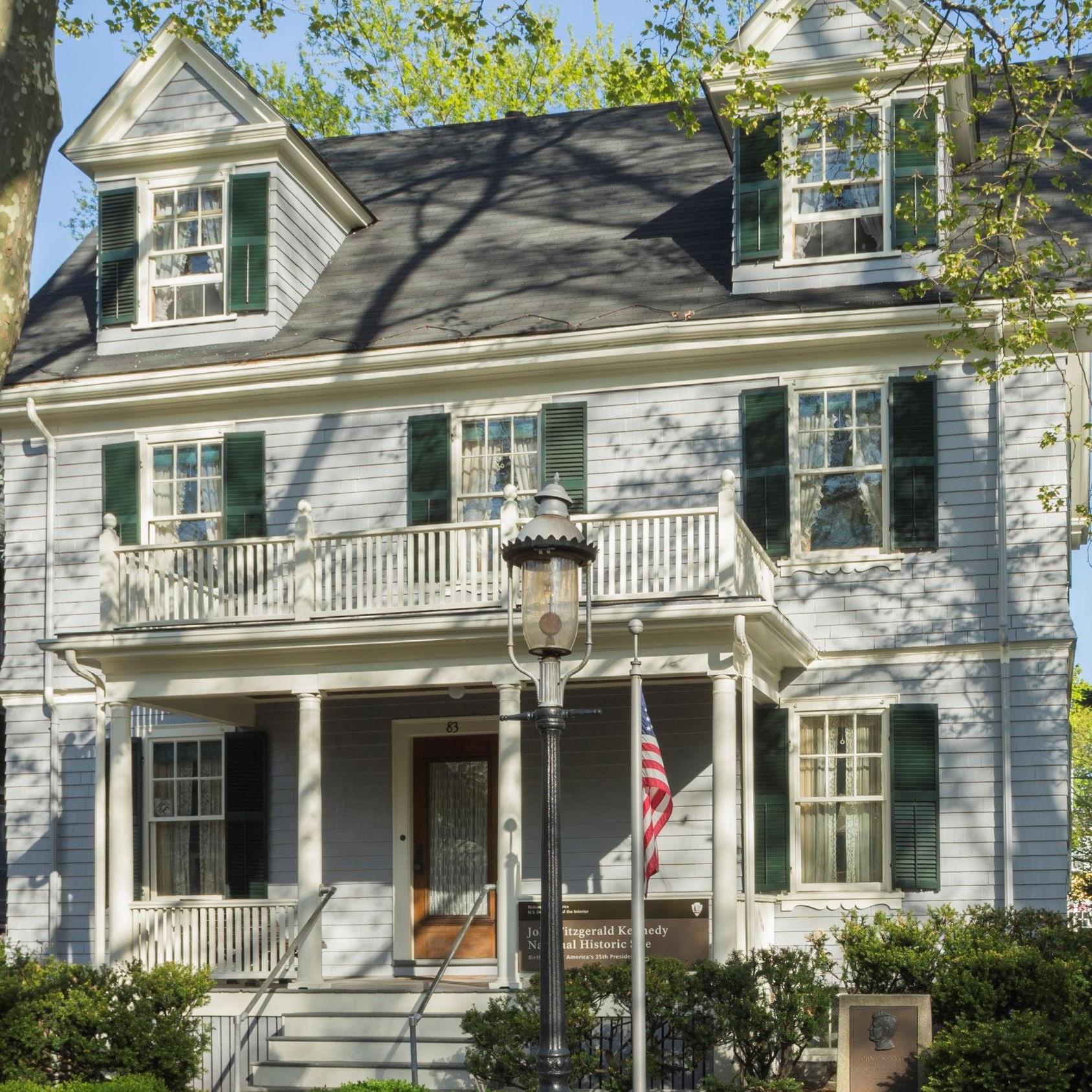 Two story gray house with two dormers in roof. Flag flying on pole in front, gas lamp on sidewalk.