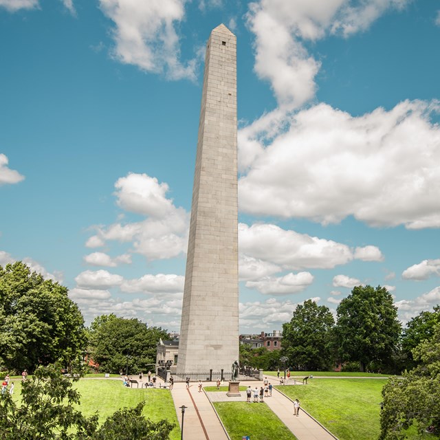 A granite obelisk stands against a bright blue sky, in the center of a manicured lawn. 