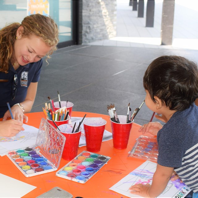 A smiling youth volunteer sits opposite an adult and child at an arts and crafts table.