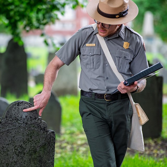 A Park Ranger stands and points at a gravestone
