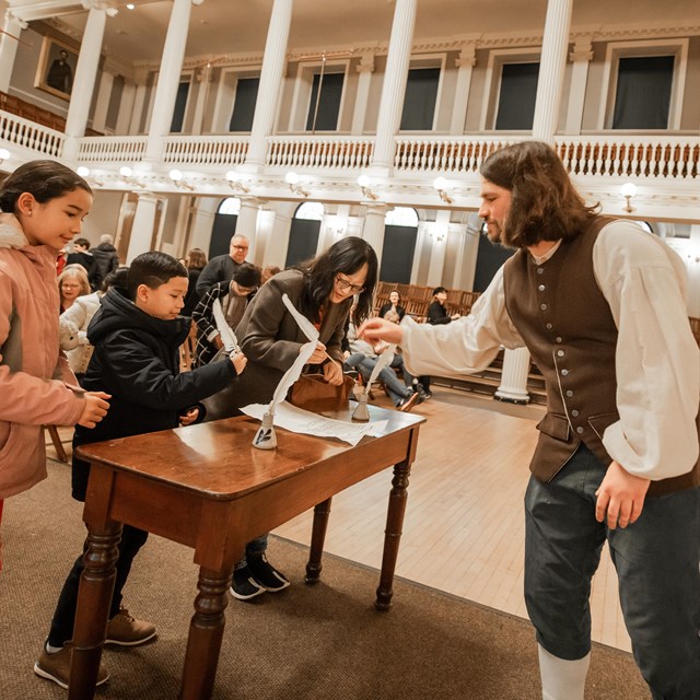 A man in colonial clothing helps kids sign a resolution