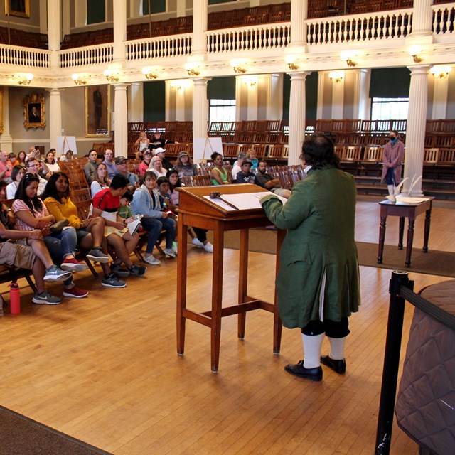 A large hall with rangers, wearing colonial period clothing, speaking to the seated audience.
