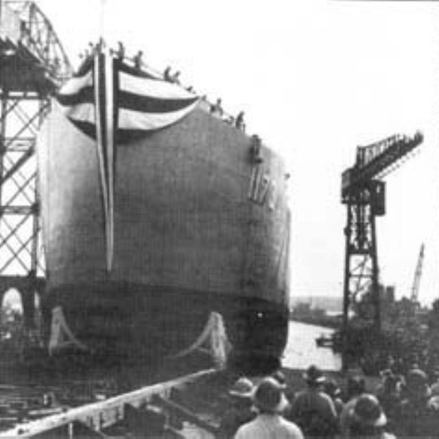 An black and white photograph of a ship being launched with onlookers in the foreground.