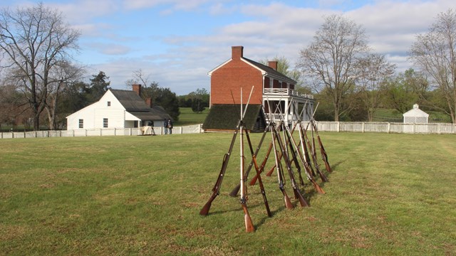 An open field with weapons stacked in a tripod formation with the Mclean House and slave quarters in