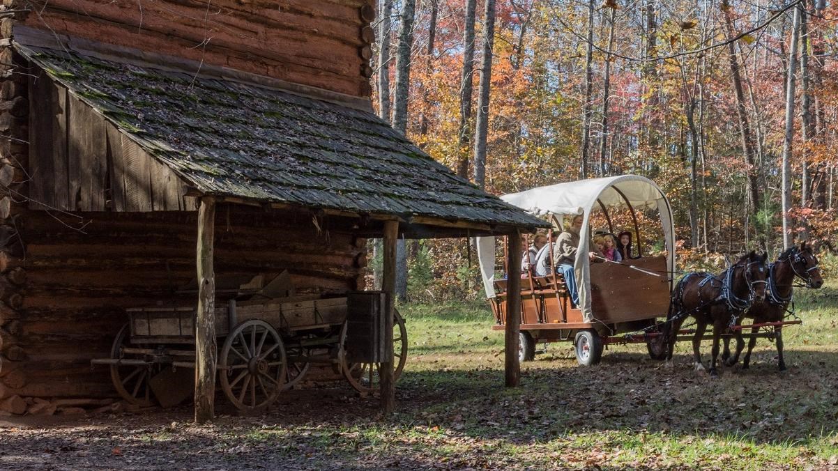 A two-horse-drawn carriage full of visitors approachs a wooden house.