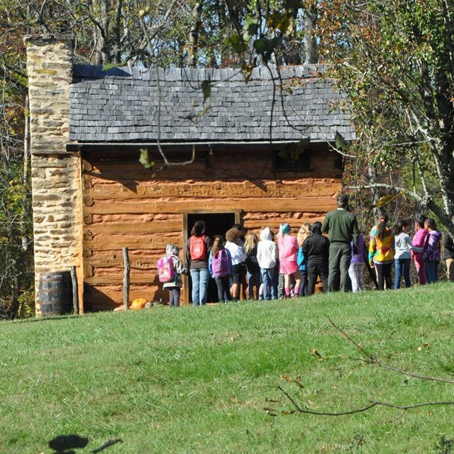 Picture of a park ranger speaking with a group of children in front of a rustic brown cabin.