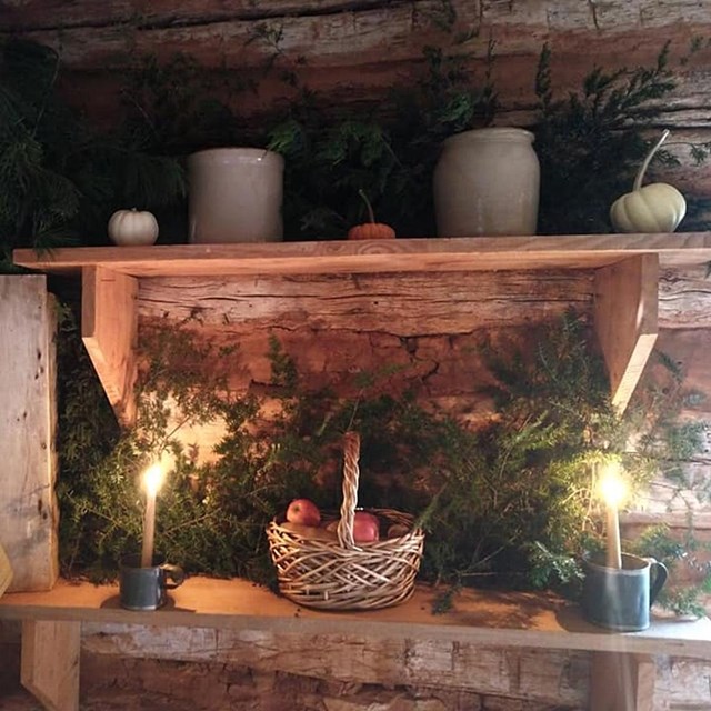 An interior view of a rustic cabin and table displaying pine boughs, lit candles and apple basket.