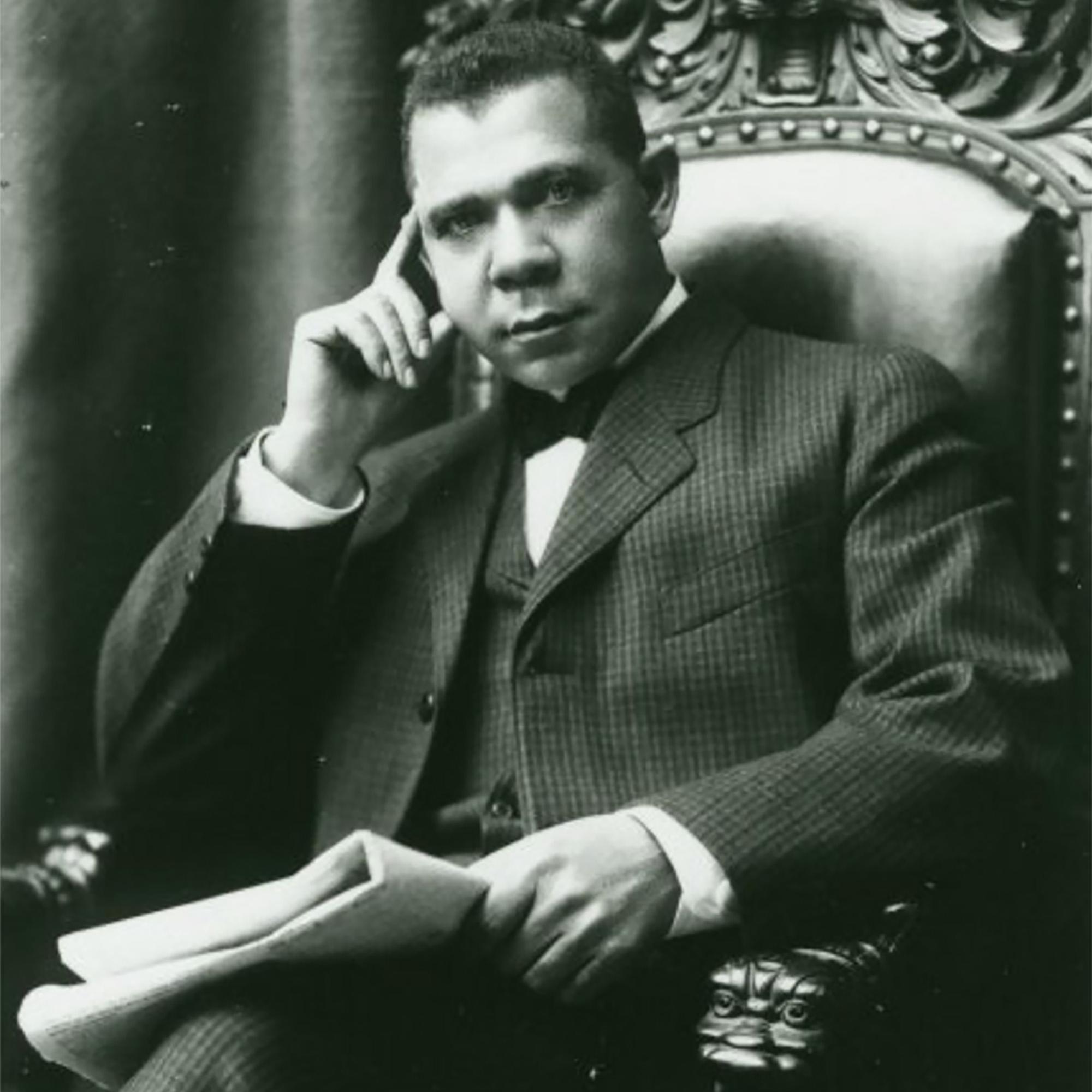A photograph of Booker T. Washington sitting in a chair with a folded newspaper in his lap. 