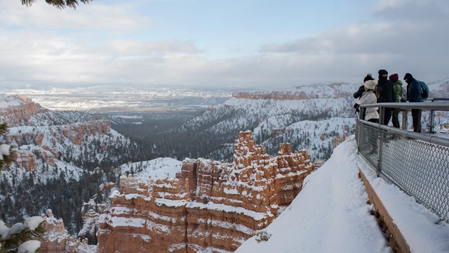 A group of people stand at an overlook looking out over snow covered red rocks.