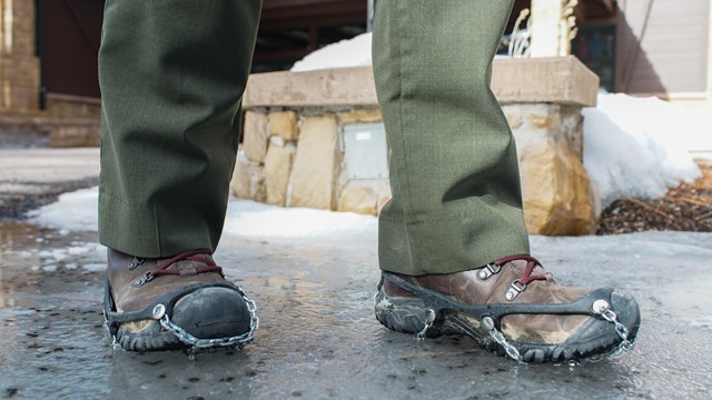 A closeup photo of two feet wearing metal traction devices over brown boots.