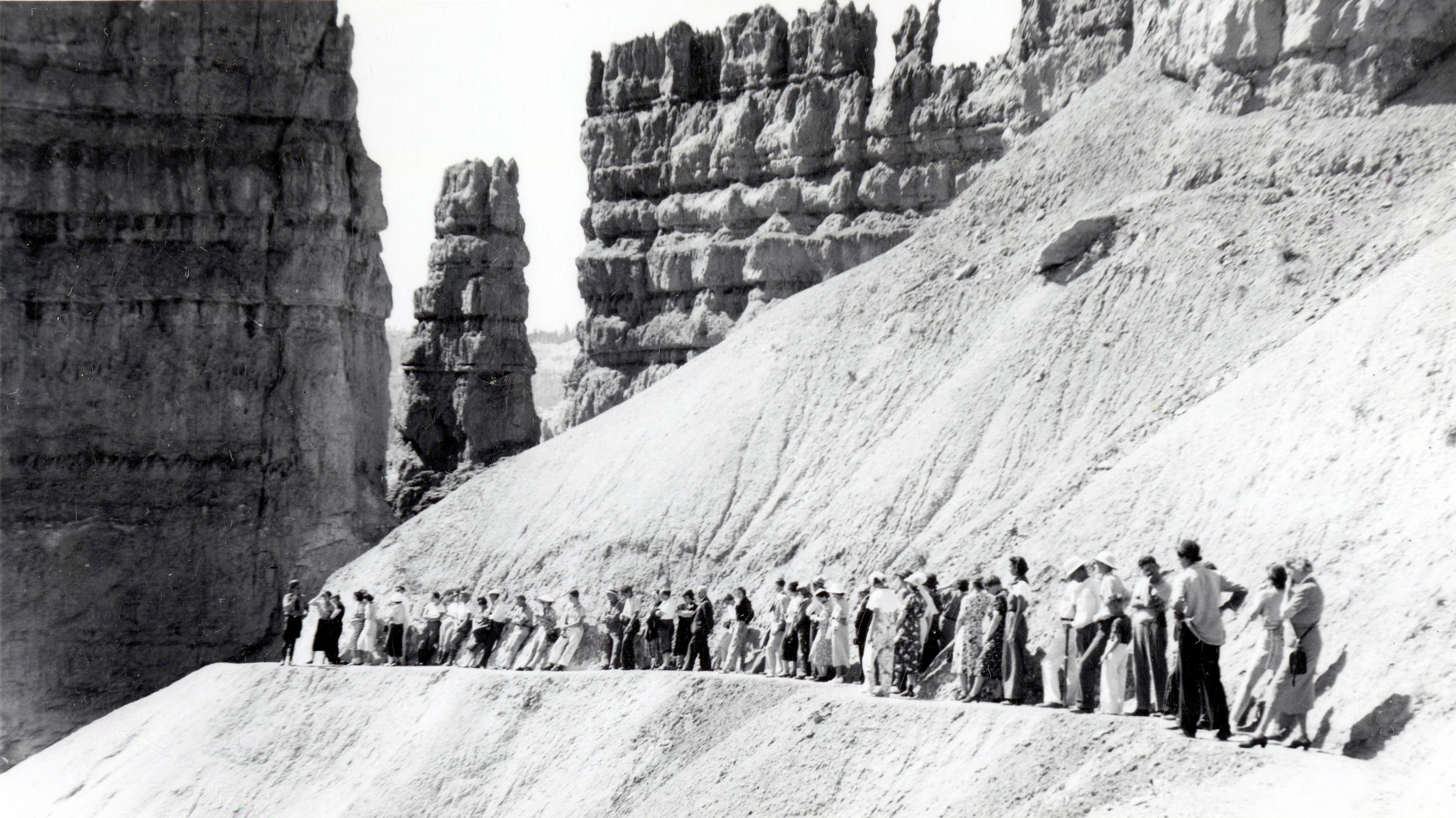 A black and white photo of a group of people along a narrow trail below tall rock spires