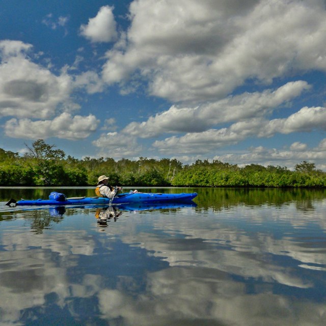 Kayaker in a wide waterway lined with trees with the clouds reflecting in the water