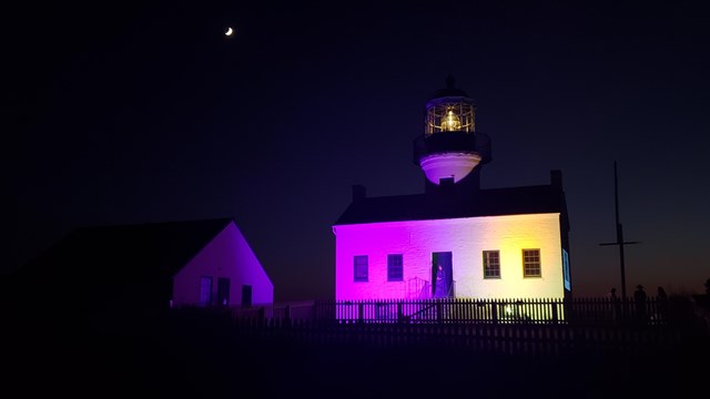 White lighthouse half lit up in purple light and other half lit with yellow lights. Yellow light in 