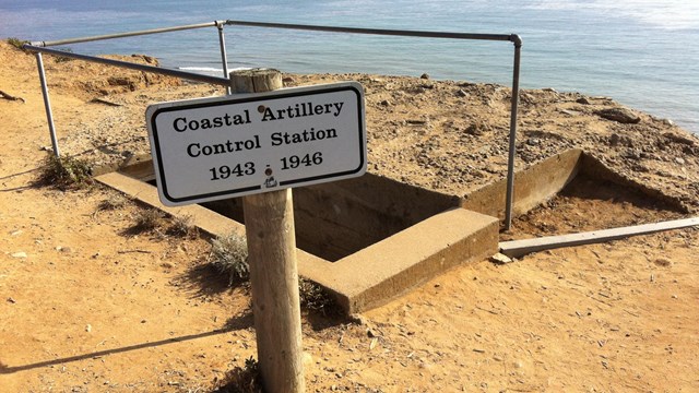 Concrete rectangle with shadow and metal rail surrounding in sandy ground. Ocean behind .