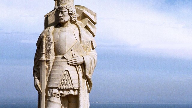 White stone statue of a man with cape. Blue ocean and blue sky with white clouds behind it.