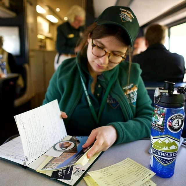 A woman with glasses sits inside of a train and looks inside a journal.