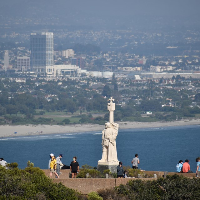 People gather around a large, white statue that stands on a lookout of a bay and green landscapes.