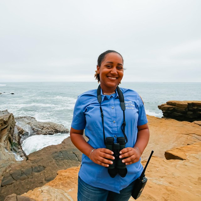 A smiling woman in a blue shirt with binoculars around her neck stands on a cliff next to the ocean.