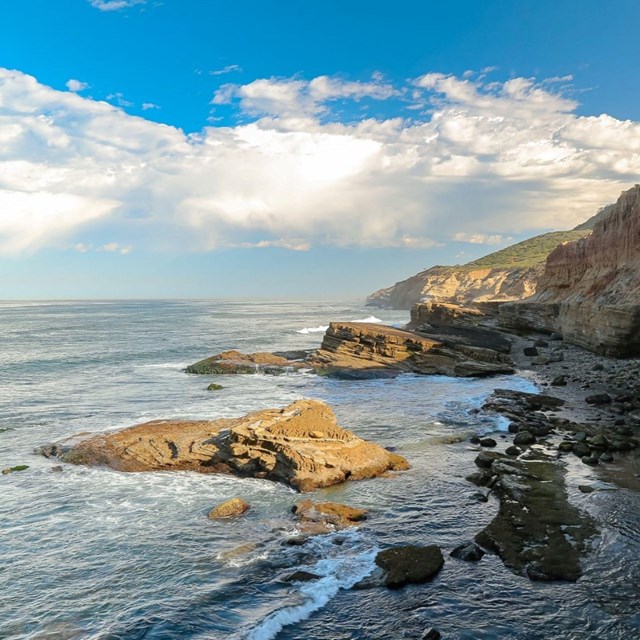Lanscape view of Cabrillo National Monument's low-tide tidepools, with blue sky, waves, and cliffs. 