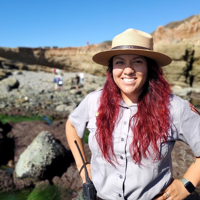 A park ranger smiles at the camera. Behind her is the rocky tidepools with bright green surfgrass. 