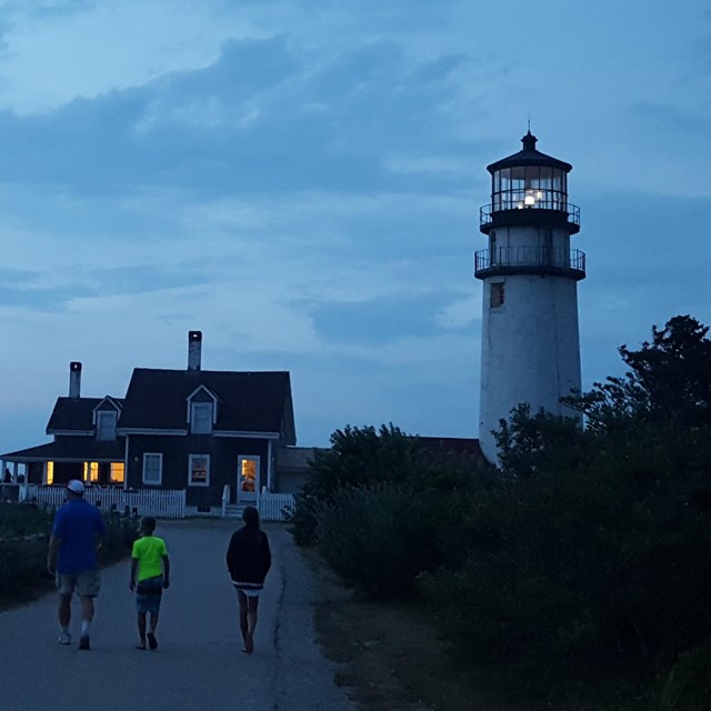 Three people walk up to a lighthouse with the tower light lit at dusk.