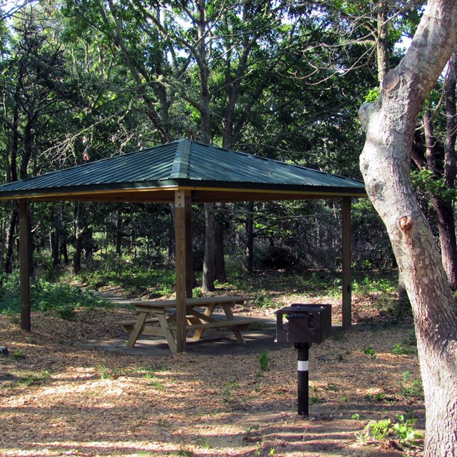 A picnic bench and barbecue grill sit in front of an open-sided wooden shelter