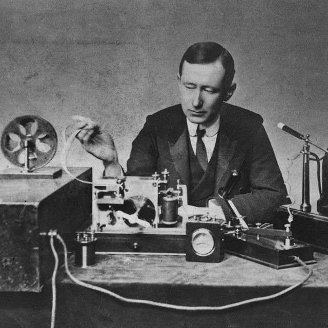 Man sits behind desk with technical gear, a transmitter and reciever.