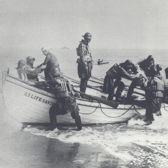 Group of men in wooden boat on beach at edge of the sea wearing cork lifevests.