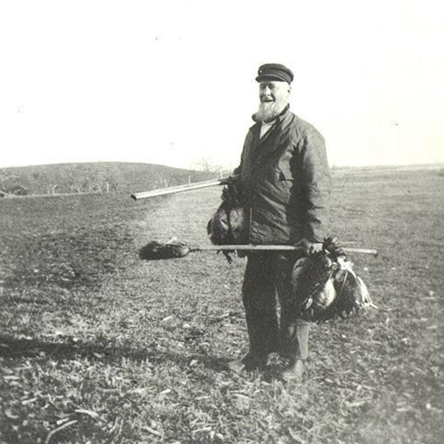 A dated black and white photograph shows a man holding small hunted game.
