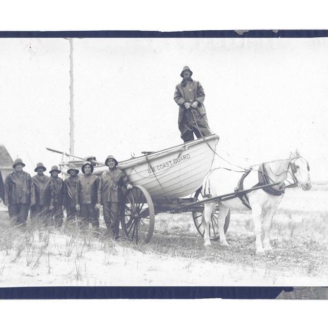 A group of men stand beside a wooden skiff boat being pulled on a cart by a horse on a beach.
