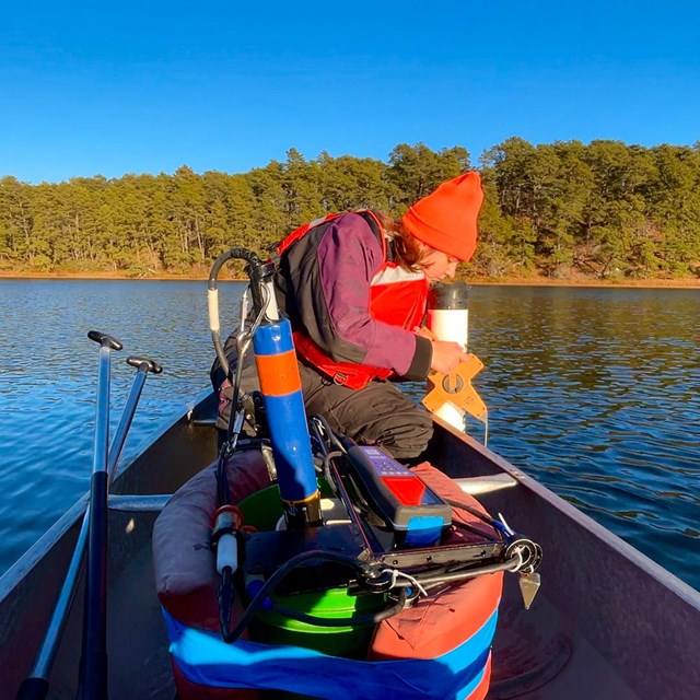 Scientist leans over edge of canoe on a pond and looks into tube at waters surface.