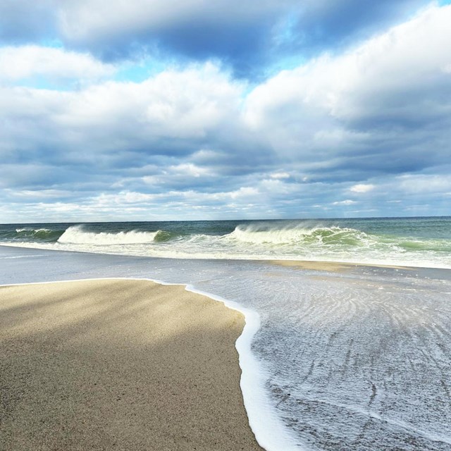 A photo of the ocean from the sand with a blue, clouded sky.