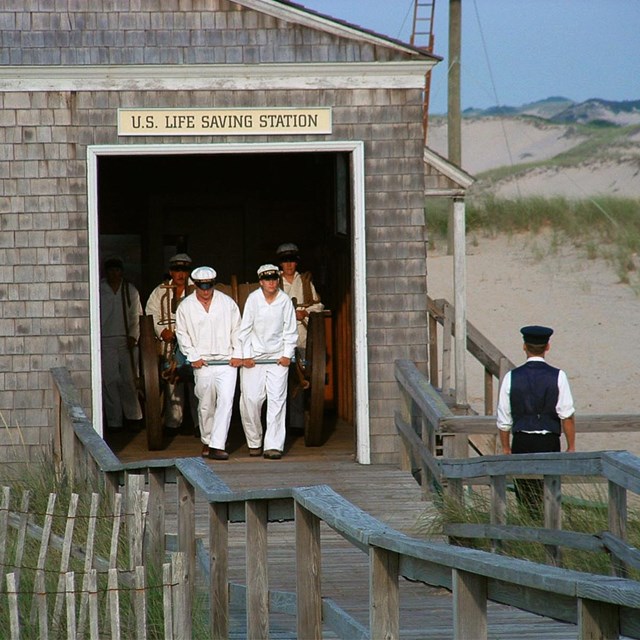 Five people dressed in Lifesavers uniforms pull a cart out of a building surrounded by beach dunes.