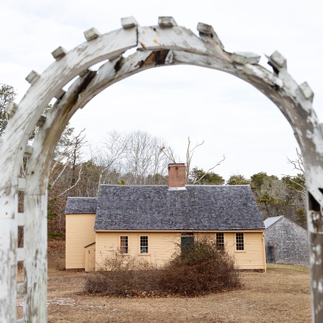 A small, yellow, one-story house can be seen through a wooden archway.