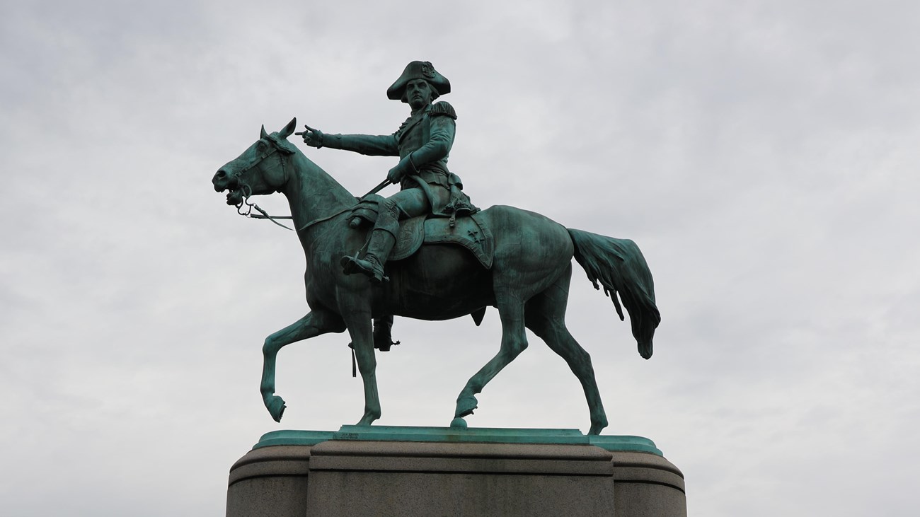 A large bronze equestrian statue of a military figure in uniform sits atop a tall stone pedestal in 