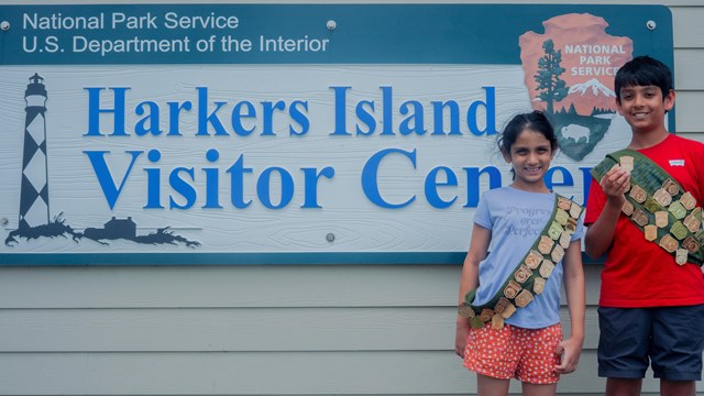 Two children stand next to the "Harkers Island Visitor Center" Sign holing their badge. 