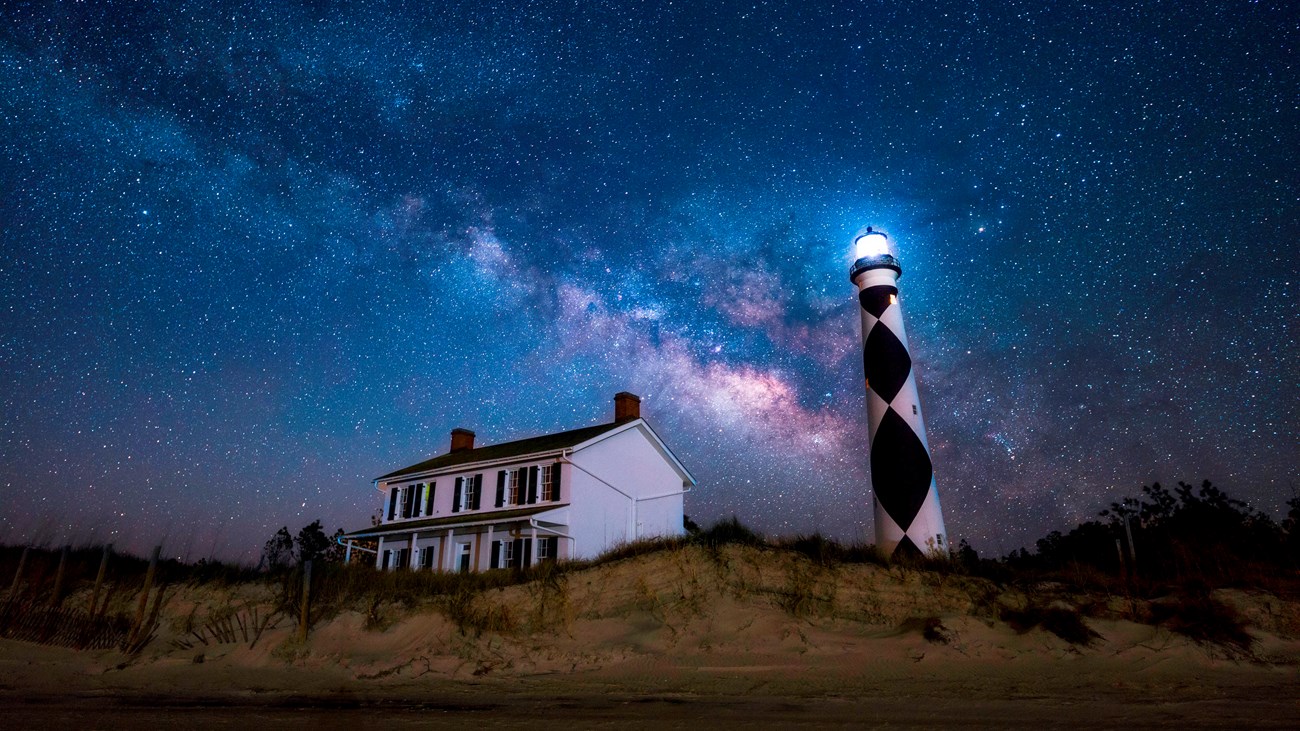 Milky Way above Lighthouse Keepers. Cape Lookout Light flashes. Sand and grass covered dunes. 