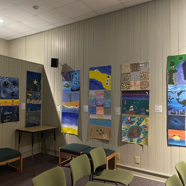 Room with painting handing on the walls. Benches and chairs visible. 