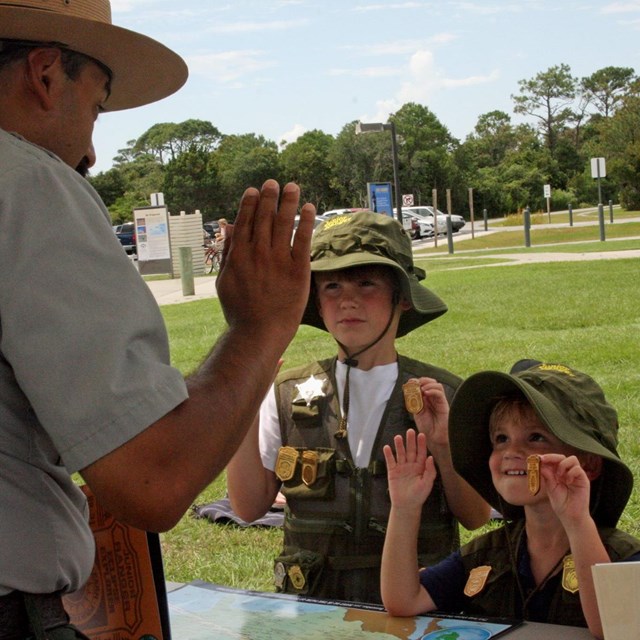 A ranger stands in front of two young children holding Junior Ranger badges. 